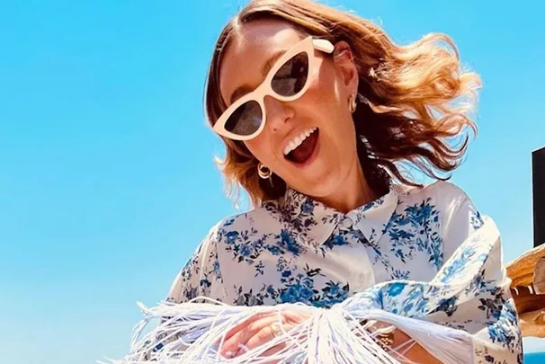 Woman in white-framed sunglasses and floral shirt smiling under a bright blue sky.