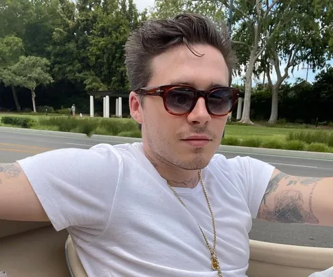 Young man with sunglasses and tattoos in a white shirt, sitting in a car with trees in the background.