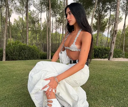 Woman in a silver top and white pants sitting on grass, with trees in the background.