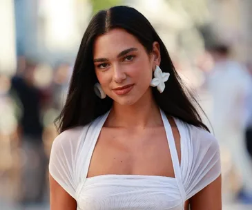 A woman with long dark hair wearing white flower earrings and a white dress, smiling outdoors.