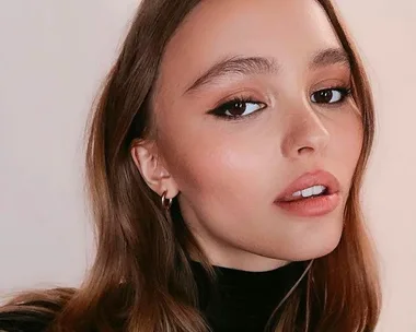 A young woman with long hair and bold eye makeup, wearing a black top, looks into the camera.