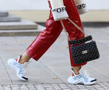 Fashionable person in red leather pants, white sneakers, and a black studded handbag, walking on a sidewalk.