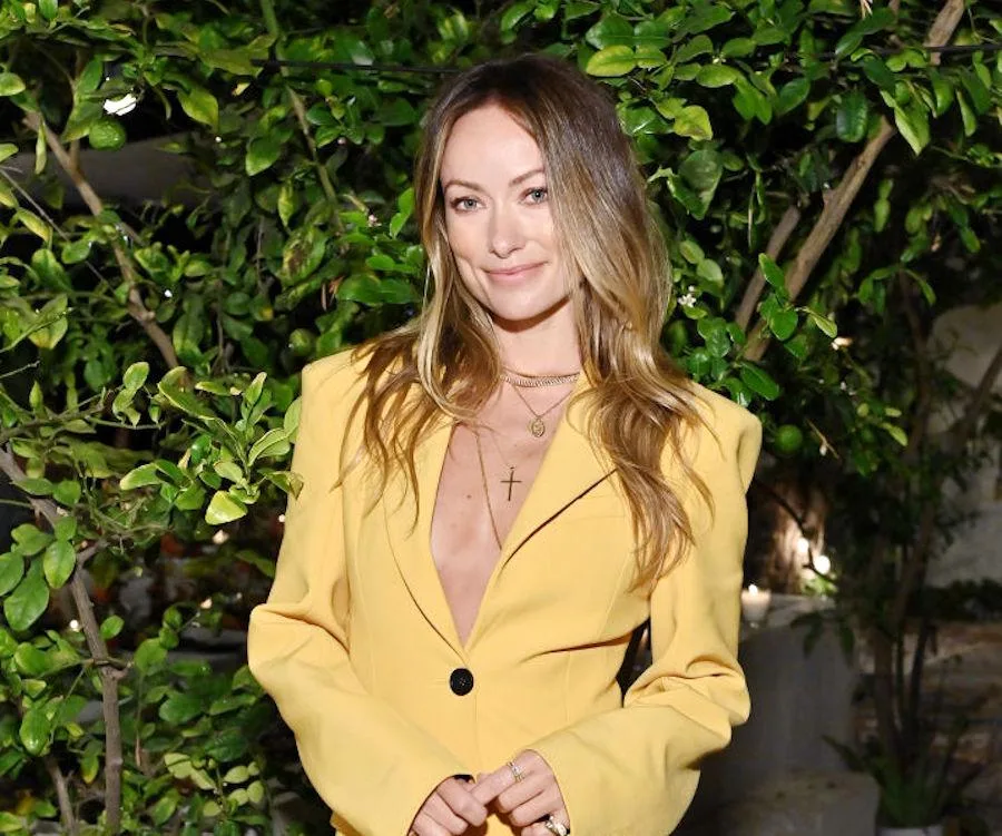 Woman in a yellow blazer standing in front of leafy greenery, smiling at the camera.