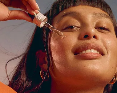 Woman applying vitamin C serum on face with a dropper, smiling, against a soft background.