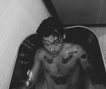 Black and white photo of a tattooed person in a bathtub, looking up, with wet hair.