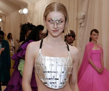 Person wearing a silver futuristic outfit with artistic face accessory at a fashion event, surrounded by other attendees.