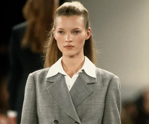 A model in a gray checkered suit and white shirt walks on a runway with a neutral expression.