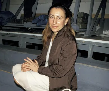 Woman seated in a brown textured jacket and white skirt, in an industrial setting.