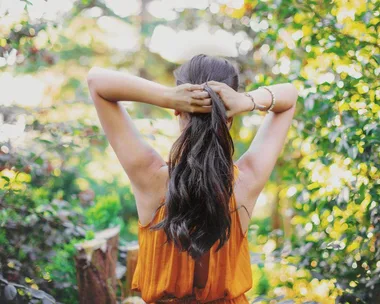 Woman in orange dress ties long, dark hair in a lush garden setting, with sunlight filtering through trees.