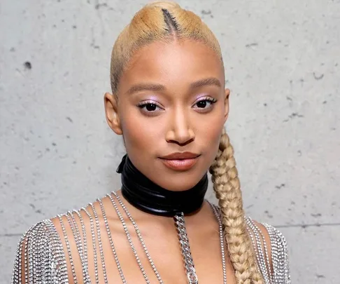 A person with blonde braided hair in a sparkly outfit and black choker, against a concrete wall.