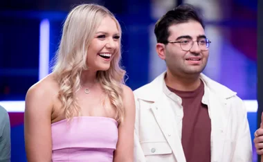 A woman in a pink dress and a man in a white jacket smile brightly, standing together in a well-lit room.