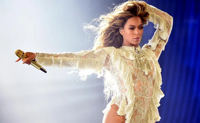 A performer in a shimmering outfit holds a gold microphone on stage with hair flowing dramatically.