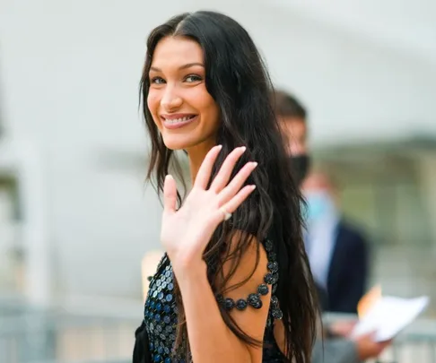 A woman with long dark hair smiling and waving, wearing an embellished dress, with blurred background.