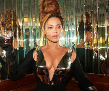 A woman with a high updo and metallic outfit holding a glass in a mirrored room.