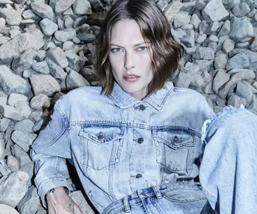 Person in denim jacket and jeans sitting on rocks, with short brown hair, looking directly at camera.