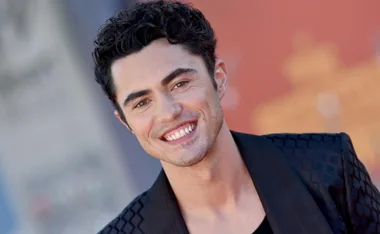 Smiling man with dark curly hair wearing a patterned black jacket, posing outdoors against a blurred background.