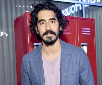 Actor in a blue blazer and pink shirt standing in front of red lockers, indoors.