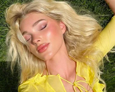 A person with long blonde hair and a yellow top lies on grass, basking in sunlight with eyes closed and a calm expression.