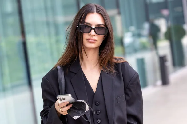 Woman in a black blazer with long hair and sunglasses, holding a phone and mask, walking outdoors.