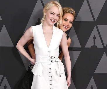 Two women smiling together at a formal event, one in a white dress, the other peeking from behind.