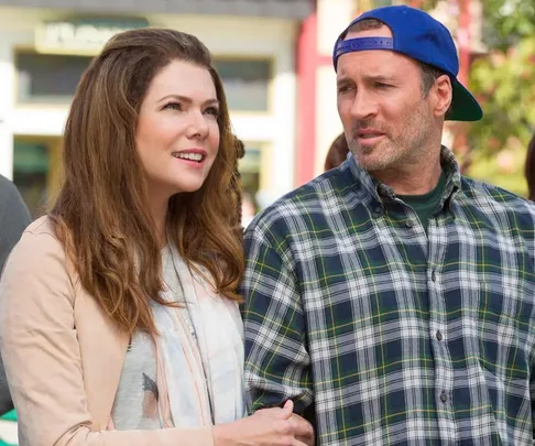 Lorelai and Luke from "Gilmore Girls" stand together, Lorelai smiling and Luke wearing a plaid shirt and backward cap.