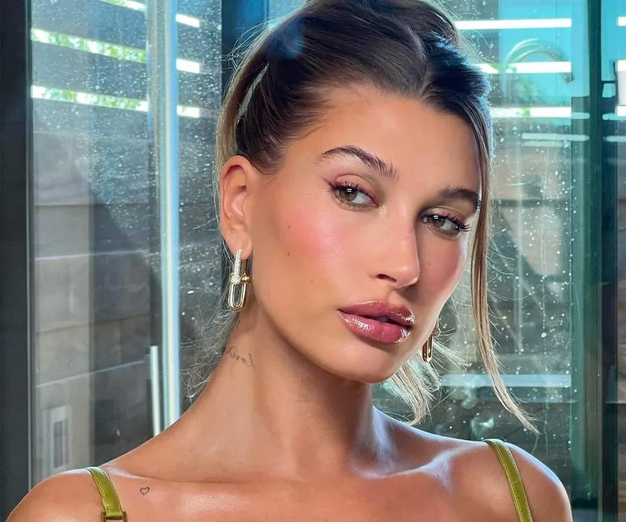 Woman with tied hair and glossy lips stands indoors, wearing hoop earrings and a tank top, looking to the side.