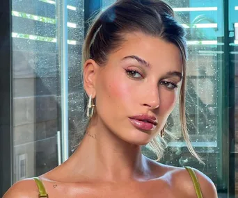 Woman with tied hair and glossy lips stands indoors, wearing hoop earrings and a tank top, looking to the side.