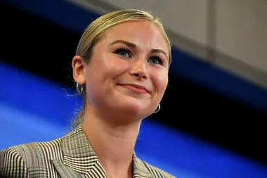 Smiling person with blonde hair and checkered blazer in front of a blue background.