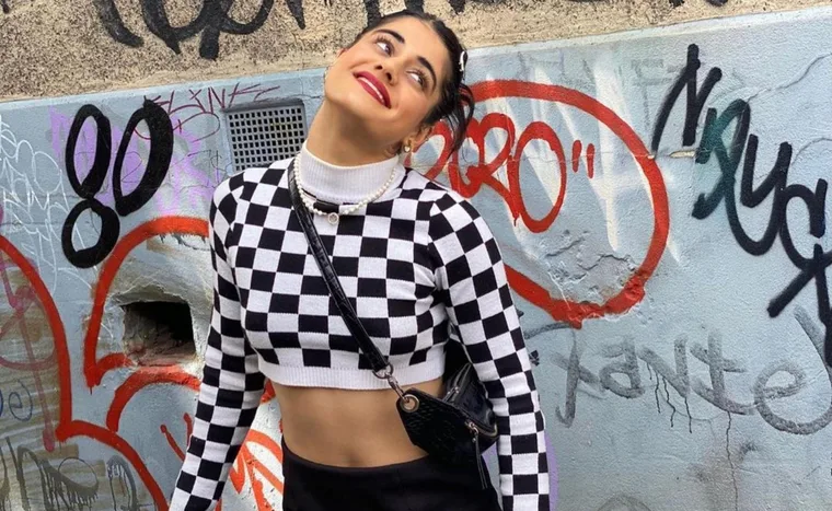 Person in black-and-white checkered crop top smiling against graffiti-covered wall.