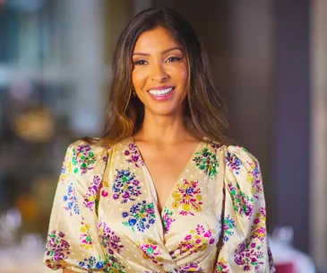 A person smiling, wearing a floral dress, standing indoors with a blurred background.