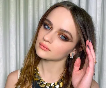 Woman with bold eyeshadow and eyeliner, wearing gold necklace, posing in front of white curtains.