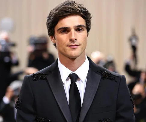 Jacob Elordi in a black suit at a formal event, with cameras in the background.