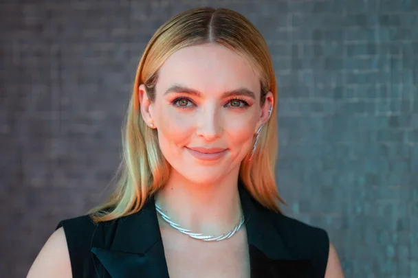 A person with long blonde hair and a black outfit, smiling against a textured background.