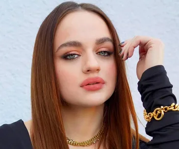 Young woman with long brown hair, wearing gold jewelry and black top, posing against a light background.