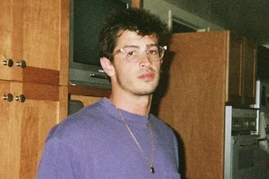 Man with glasses and a purple shirt standing in a wooden kitchen setting.