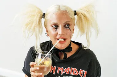 Blonde woman with dramatic eye makeup, pigtails, wearing band shirt, sipping drink through straw.