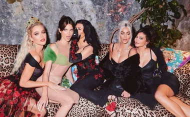 Group of five women in glamorous outfits sitting on a leopard print couch, posing for a photo.