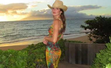 A woman in a colorful dress and cowboy hat stands on a beach at sunset, with the ocean and sky in the background.