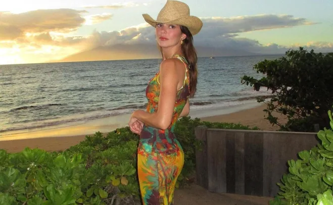 A woman in a colorful dress and cowboy hat stands on a beach at sunset, with the ocean and sky in the background.