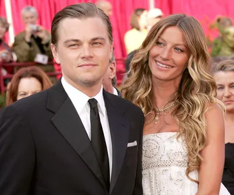 A man in a black suit and tie stands beside a woman in a white dress with long wavy hair. They are outdoors at an event.