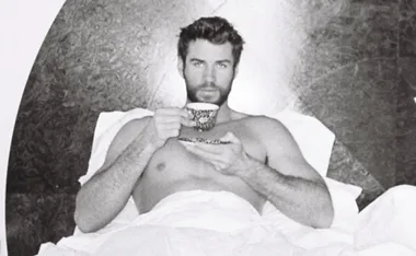 Shirtless man with beard in bed, holding a cup and saucer, black and white image.