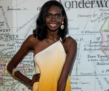 Woman in a sleeveless yellow dress smiling, stands in front of a vintage world map background.