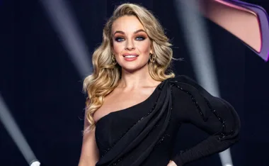 A woman in a glamorous black dress poses confidently on stage with styled blonde hair, smiling for the camera.