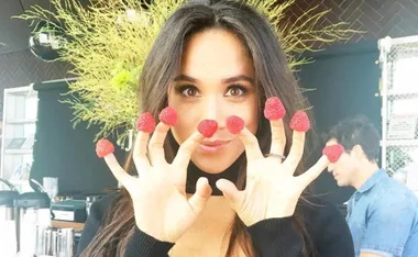 Woman holding raspberries on all her fingertips, playfully smiling at the camera, with foliage in the background.