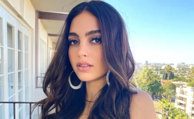 Portrait of a woman with long brown hair and hoop earrings standing on a balcony with a cityscape in the background.