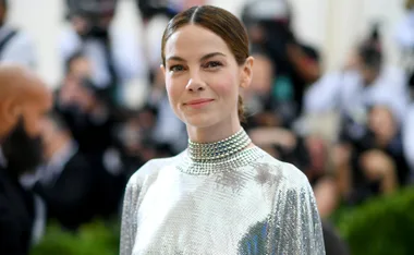 A woman in a silver, bejeweled dress smiles at an event, with a blurred crowd and cameras in the background.