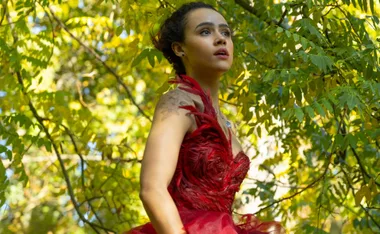 Woman in a red feathered dress under trees, surrounded by sunlight.