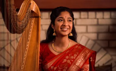 Young woman in a red sari smiling by a harp, likely from "Never Have I Ever" Season 4.