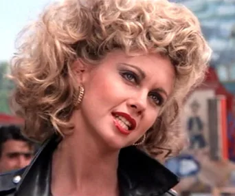 Olivia Newton-John as Sandy in "Grease," wearing a black leather jacket and curly blond hair, with a confident expression.