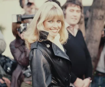 Blonde woman in a black leather jacket smiling at a public event, with photographers in the background.